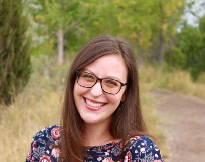 Michelle Garcia, licensed marriage and family therapist, registered play therapist, smiling, wearing glasses and a dark blue shirt with white, pink, and red flowers, standing outdoors with cottonwood trees and grass in the background