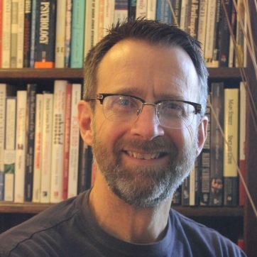 Dr. Russell Thye, smiling, wearing glasses, standing in front of a bookcase