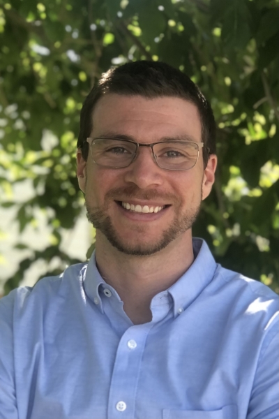 Nicholas Terry, licensed professional counselor, in the shade of a tree, smiling, wearing glasses and a light blue button-up shirt