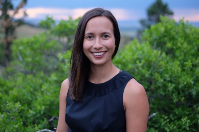 Alisha Johnson, licensed clinical social worker, wearing a dark blue sleeveless top, smiling, standing outdoors with bushes/trees and a beautiful sky in the background.