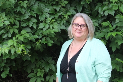 A woman in a pale green sweater standing in front of green bushes.