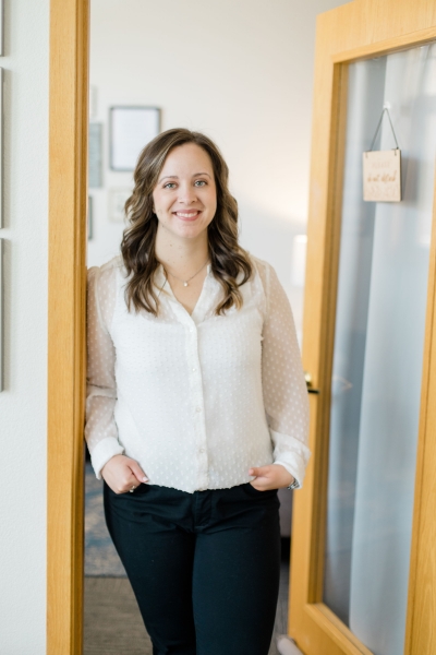 Samantha Ricard, licensed marriage and family therapist, smiling, wearing a white shirt and black pants, stranding in the doorway to her office.