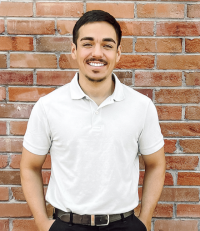 Jorge Rubio, licensed professional counselor candidate, smiling, wearing dark pants and a white, short-sleeve polo shirt, standing in front of a brick wall