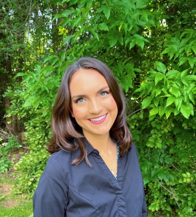 Julia Schafer, clinical social work candidate, smiling, standing next to a leafy green tree, wearing a blue shirt