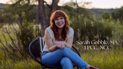Sarah Gobble, licensed professional counselor candidate, smiling, sitting outdoors in a chair, with a tree and bushes in the background