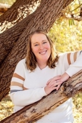 Emily O'Rourke-Mills, licensed marriage and family therapist, smiling, standing outdoors leaning against a tree, wearing a white, long-sleeved shirt with brown stripes on the sleeves