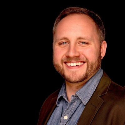 Cody Watts, licensed professional counselor candidate, smiling, wearing a blue button-up shirt and dark brown jacket against a black background