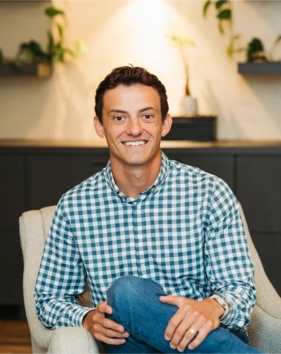 Kaine Fredrickson, licensed professional counselor candidate, smiling, sitting in a chair, wearing jeans and a blue and white plaid shirt.