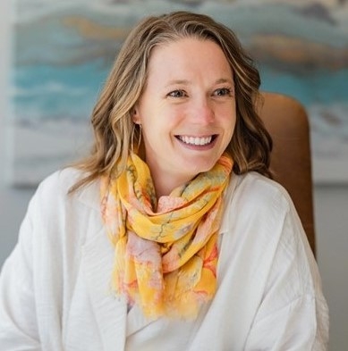 Abbey Schneider, licensed marriage and family therapist, smiling, sitting in a chair and wearing a white top with a colorful yellow scarf