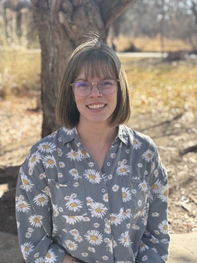 Clair Witt, licensed professional counseor candidate, smiling, wearing glasses and a gray shirt with white and yellow flowers, with a tree in the background