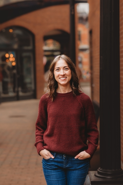 Angie Bartleson, licensed marriage and family therapist, licensed addiction counselor, smiling, wearing jeans and a maroon sweater, standing with hands in her pockets, with brick buildings in the background