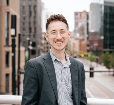 Drew Feldbaum, licensed clinical social worker, smiling, wearing glasses, a striped, button-up shirt and a jacket, with a cityscape in the background