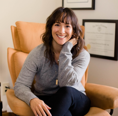 Dori Lewis, licensed professional counselor, smiling, sitting in a chair in her office, wearing dark blue pants and a light gray, long-sleeved top.