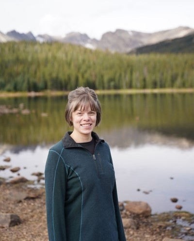 Claire Witt, licensed professional counselor candidate, smiling, with short hair, with a lake and mountains in the background.