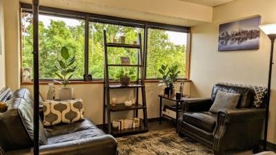 Office with large windows, sofa and chair, potted plants, and display shelf..
