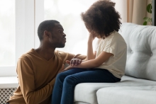 Father sitting on the floor consoling his young daughter who is sitting on a couch.