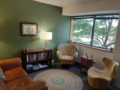 Office space with a sofa, comfortable chairs, windows, a small bookcase, circular area rug, and floor lamp.