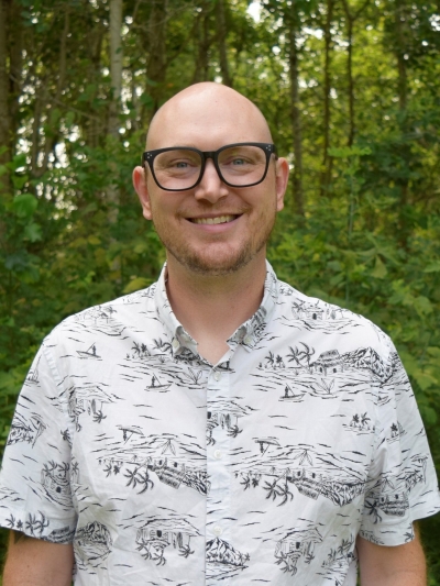 Aaron Schuler, licensed professional counselor candidate, smiling, wearing dark-framed glasses and a short-sleeved, white shirt, with black designs 