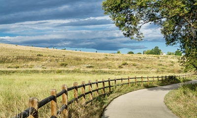 A paved path next to a split-rail fence, with a tree and view of an open field of prairie grass.