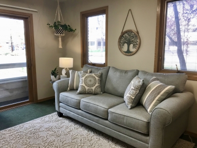 Office with windows, area rug, table lamp, potted plants, and sofa with pillows. 