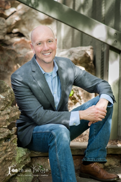 John Kinnaird, licensed clinical social worker, owner and therapist at Mindful Transitions, smiling, sitting outdoors, wearing jeans, a gray suit jacket, and light blue button-up shirt.