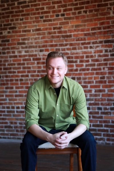 Zach Engler, licensed professional counselor, smiling, sitting on a chair, wearing a green button-up shirt and dark pants