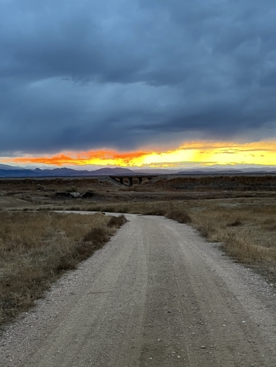View along a dirt road with a cloudy sky and sunset visible between the foothills and clouds