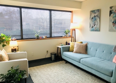 A therapy room at Bloom Counseling and Nutrition, with light blue sofa, a white chair, floor lamp, and windows